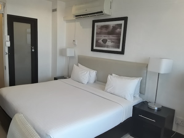 Cama bem arrumada com lençóis brancos em quarto de hotel. Há uma moldura com um quadro na parede acima da cama, um abajur e uma pequena gaveta sob a cama.
