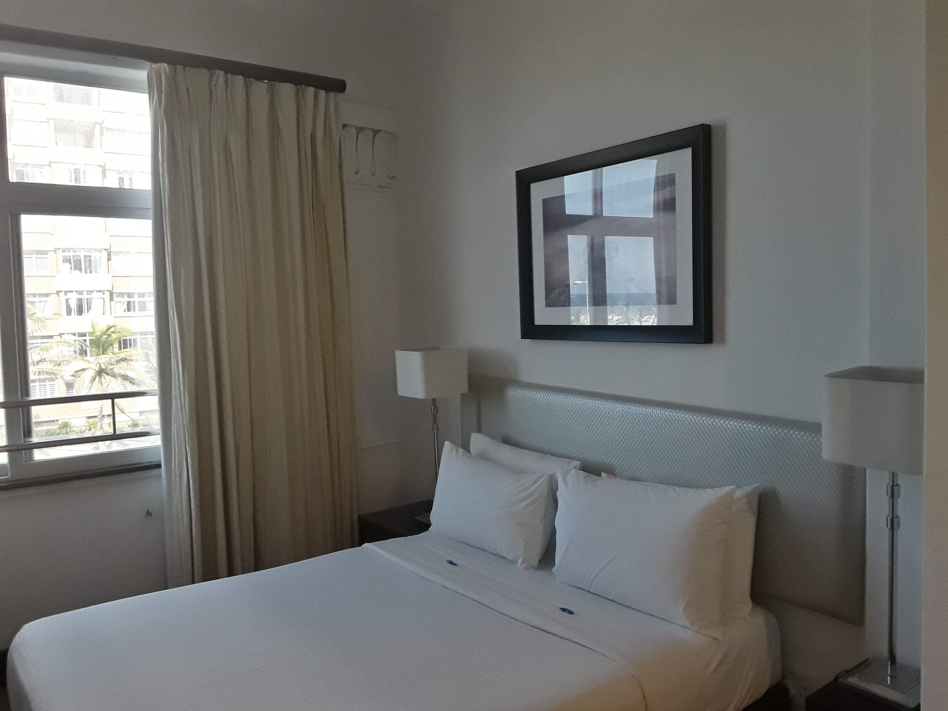 A hotel room with a bed, a window with curtains, a framed picture on the wall, and a nightstand with a lamp.