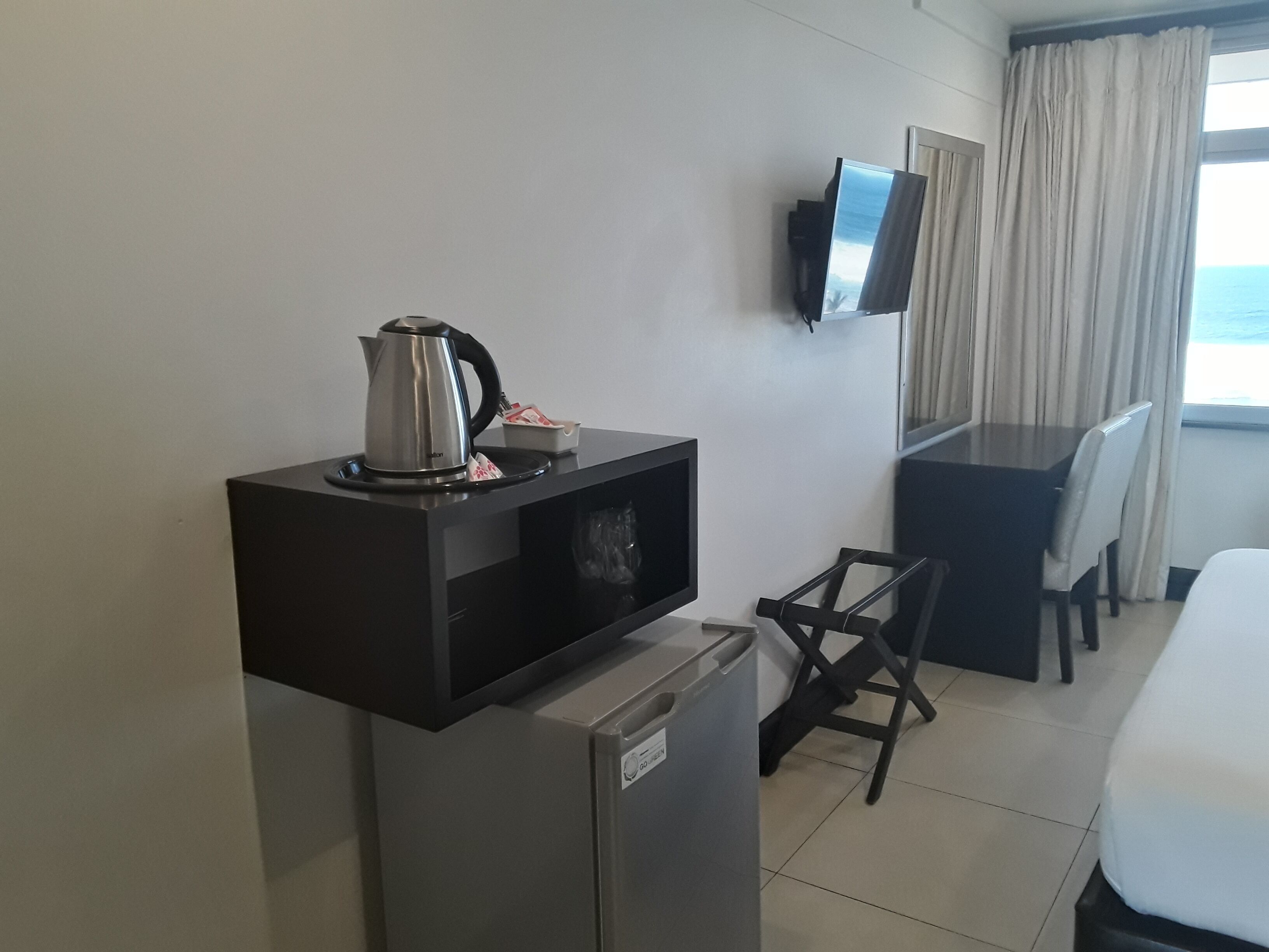 A compact hotel room with a kitchenette, a flat-screen TV mounted on the wall, a dining area with a table and chairs, and a large window offering a view of the sea.