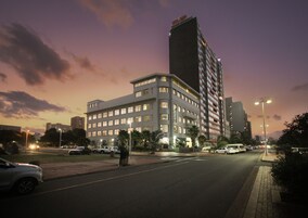 Un hotel de varios pisos con diseño moderno, ubicado en una esquina destacada, con una entrada amplia y un área de estacionamiento frente a él.