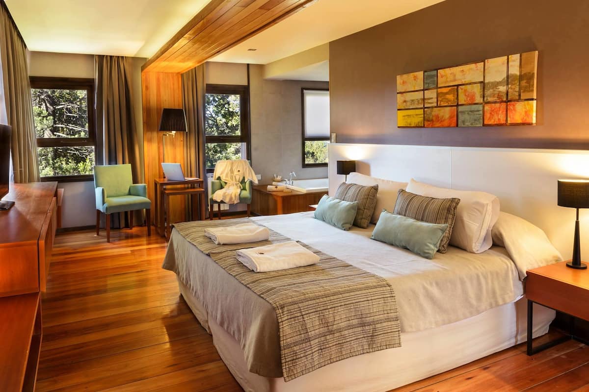 A modern hotel room with a large bed, a desk, a chair, a small table, and a window with curtains.
