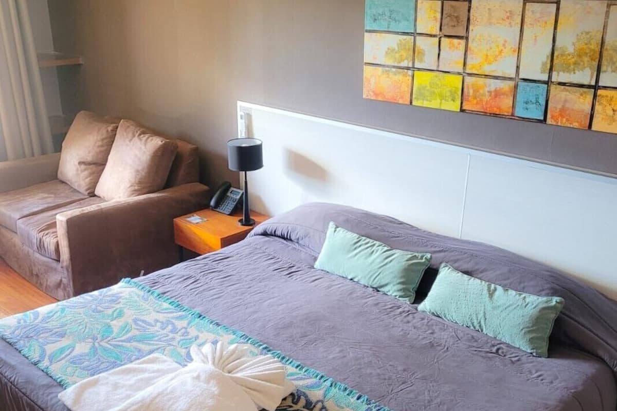 A bedroom with a large bed, a chair, a bedside table with a lamp, and a wall with a colorful artwork.