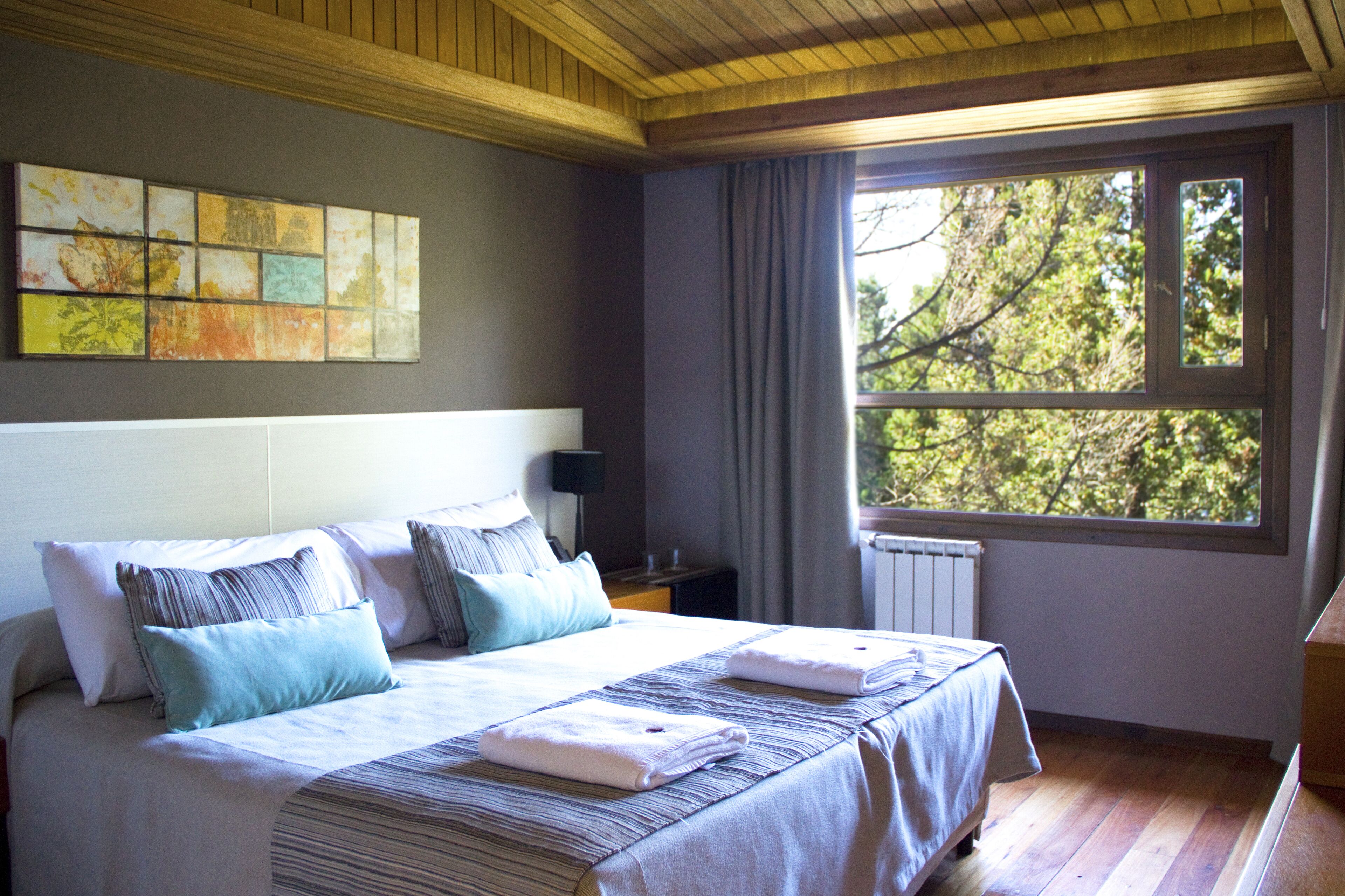 Quarto com cama grande, janela com vista para árvores e parede com obra de arte colorida.