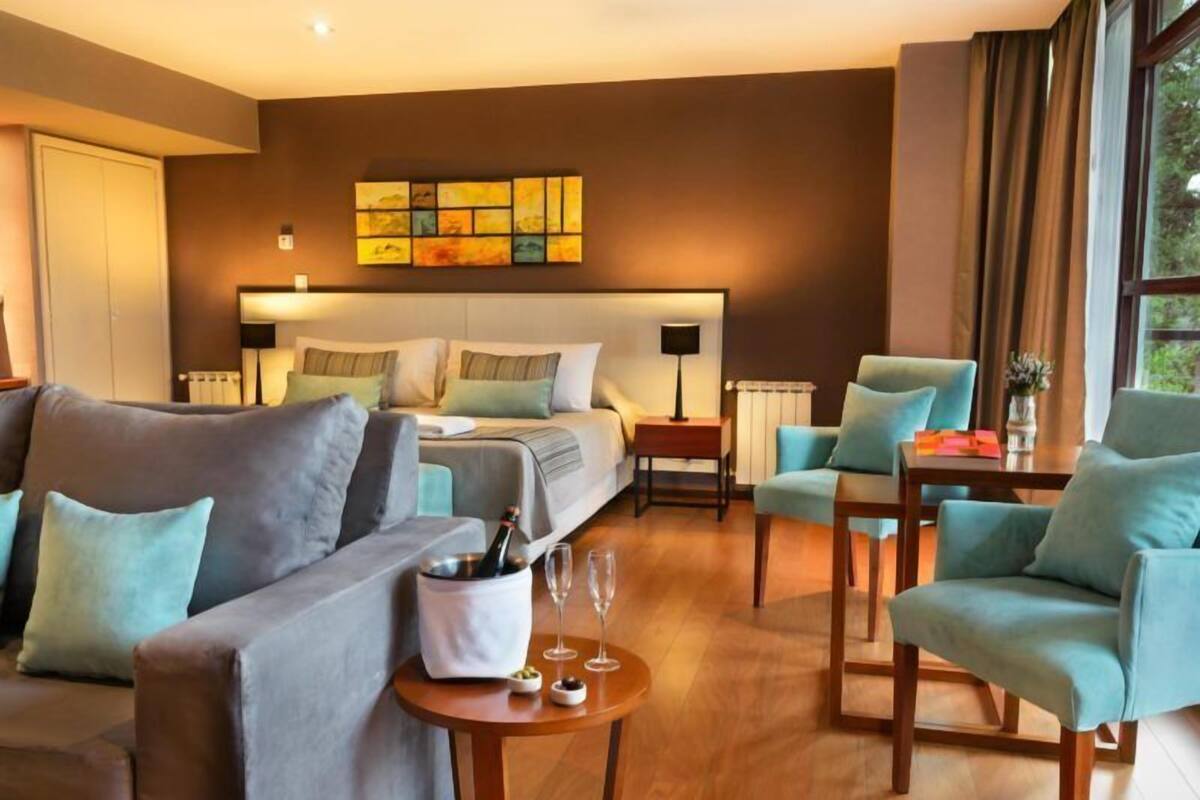 A modern hotel room with a bed, a sofa, a dining table, and a view of the outdoors.