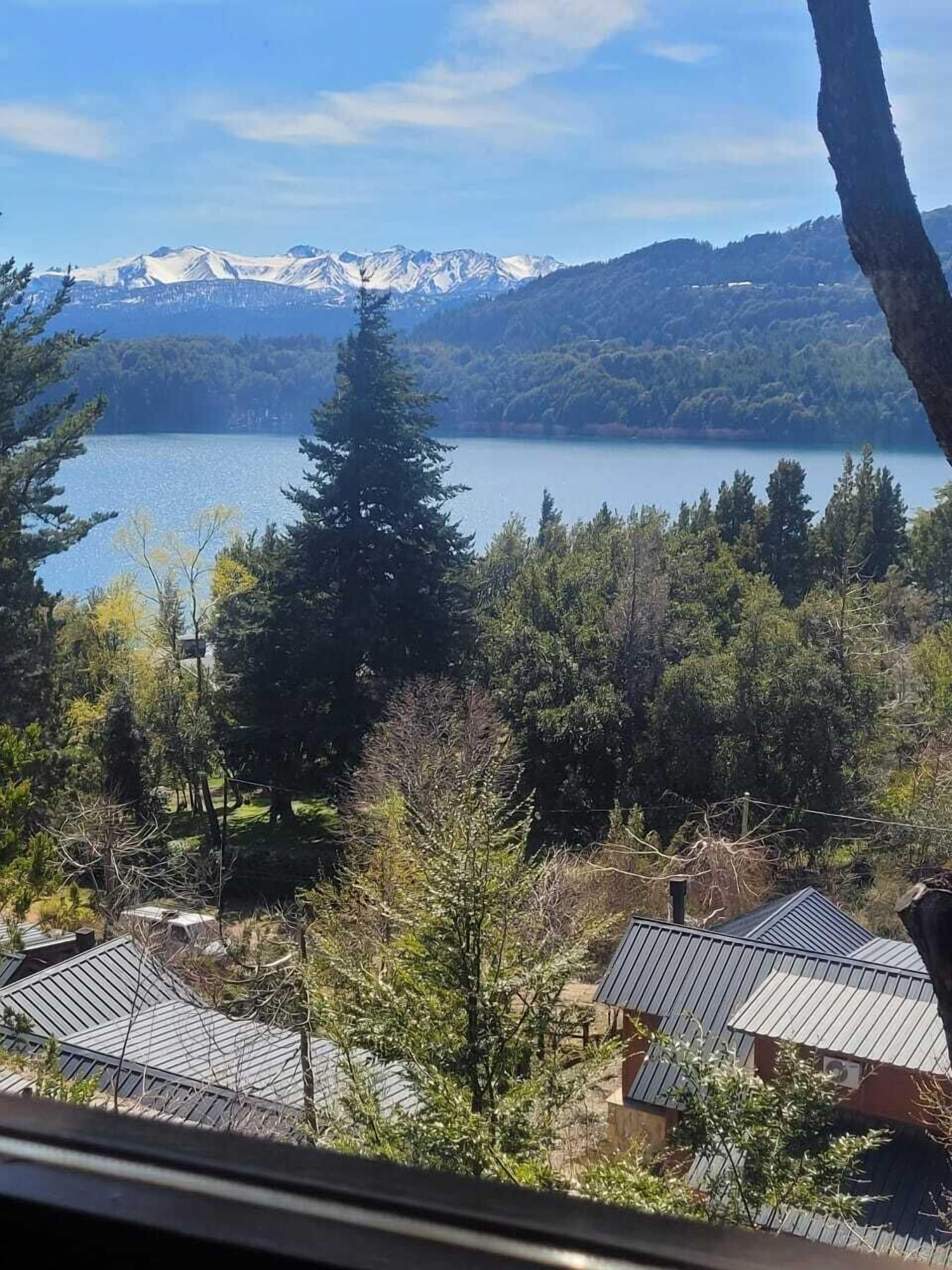 Ein See mit Bergen im Hintergrund, umgeben von Bäumen und Gebäuden mit Metalldächern.