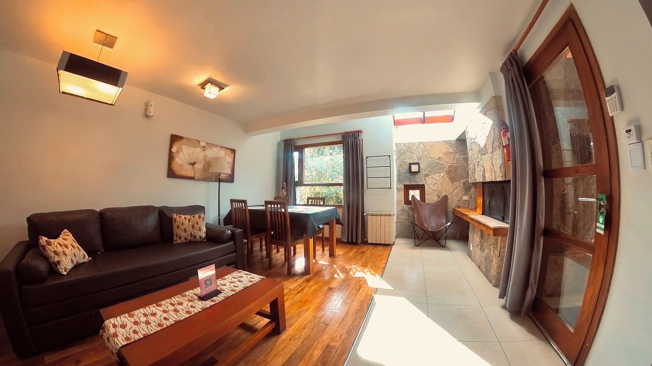 A living room with a sofa, coffee table, and a dining area with chairs and a table. There is a fireplace with a stone surround and a window with curtains.