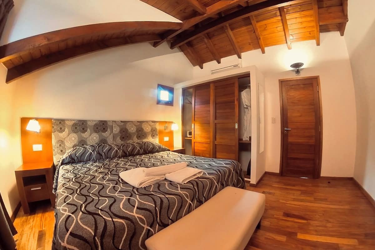 A bedroom with a bed, bedside tables, a bench, wooden ceiling beams, and a wardrobe.
