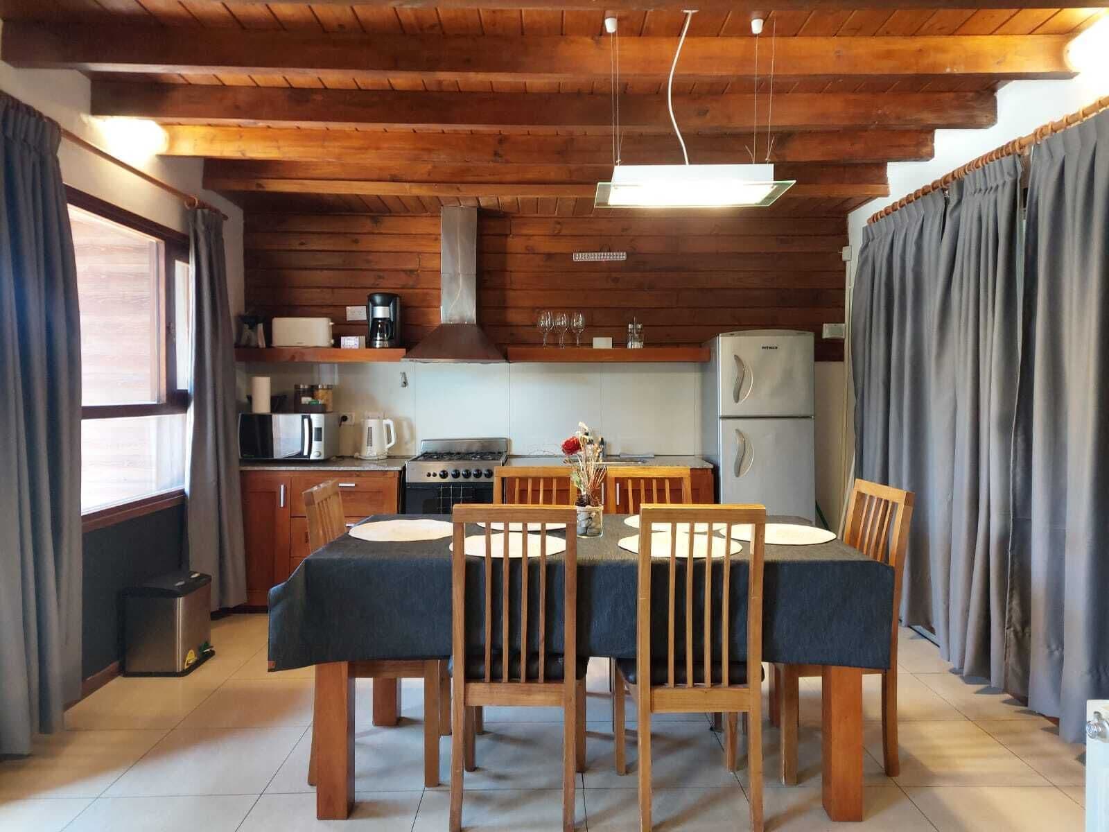 Una cocina acogedora con armarios de madera, una estufa y un refrigerador. Hay una mesa de comedor con sillas y una ventana con cortinas.