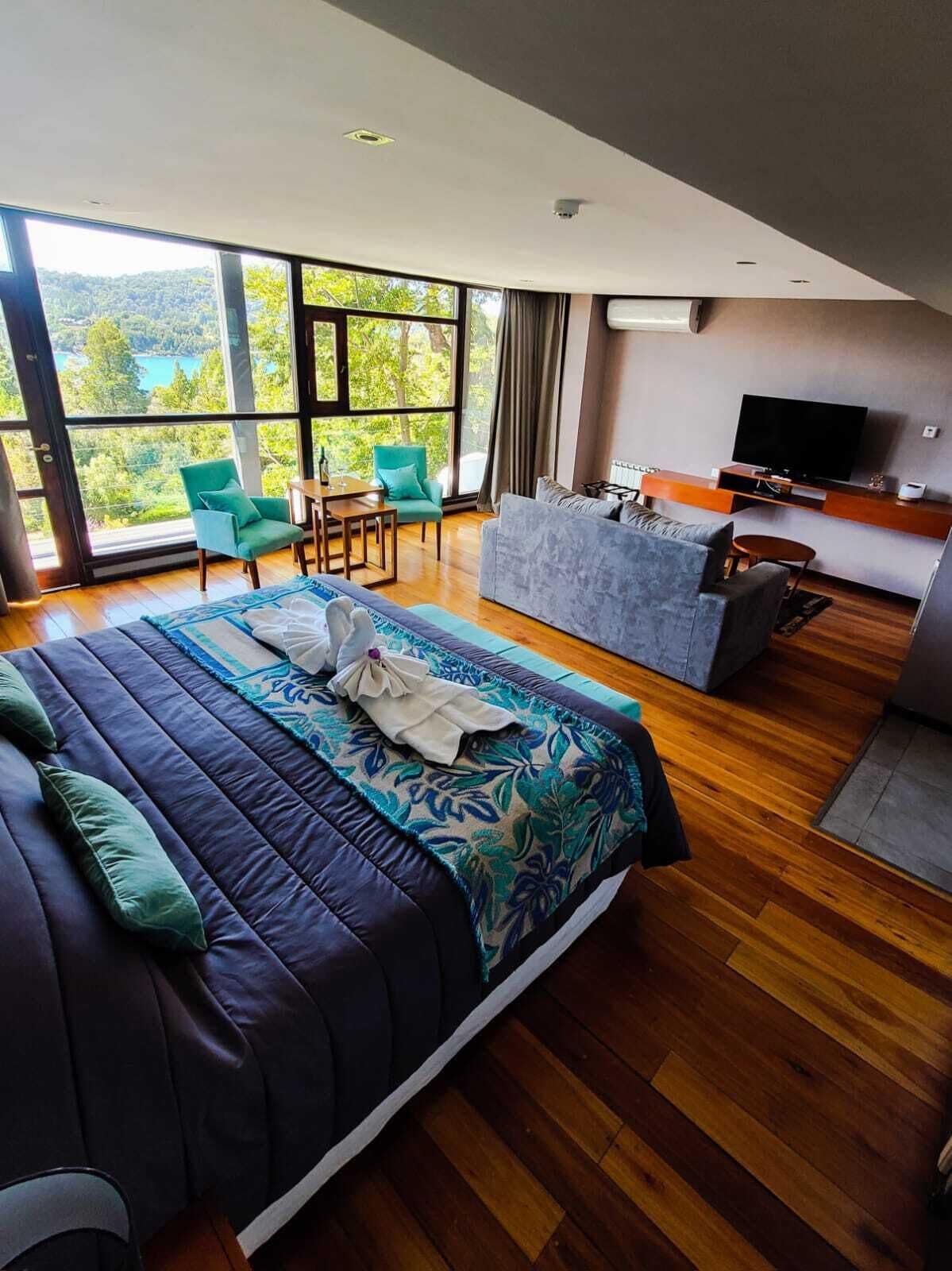 A modern hotel room with a large bed, a sofa, a TV, and a balcony with a view of the outdoors.