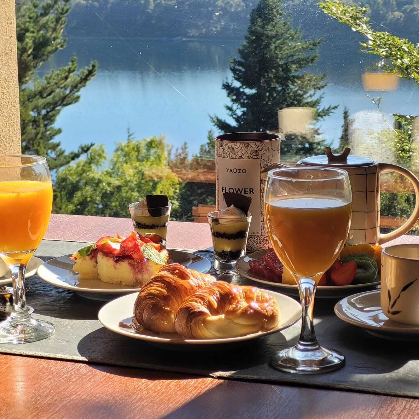 Ein Frühstückstisch mit Blick auf einen idyllischen See, mit einem Croissant, Obst und einem Getränk.