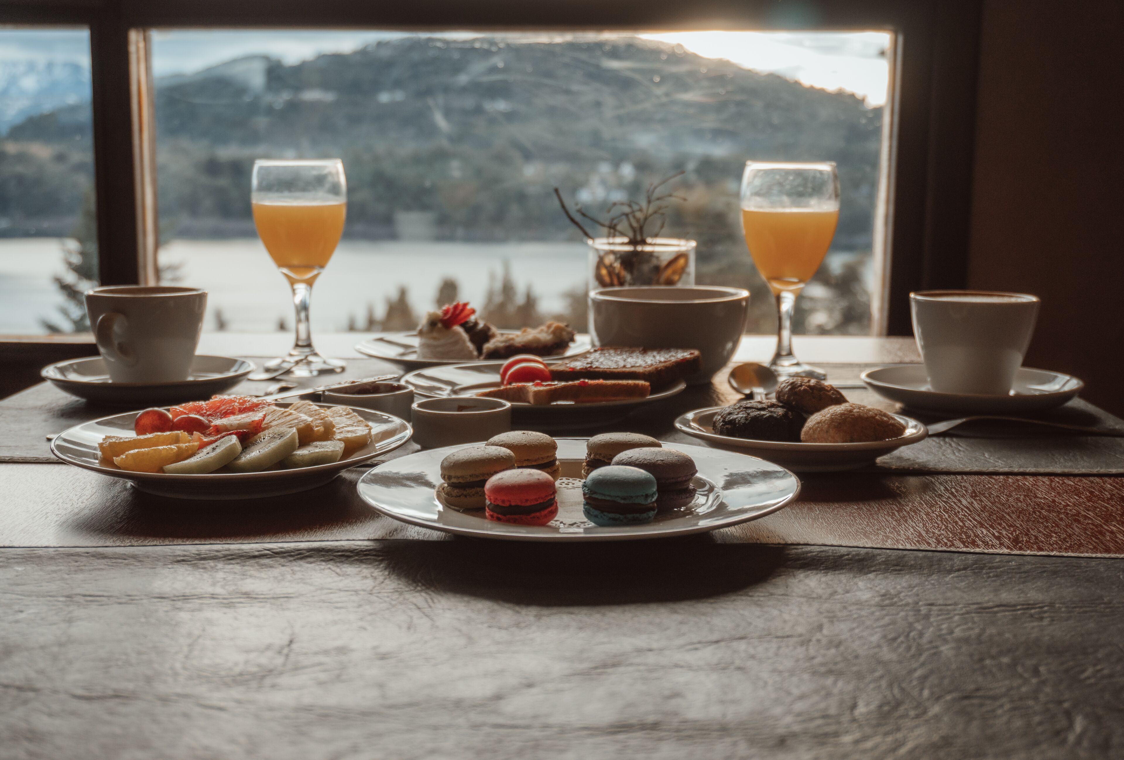 Une table garnie de diverses pâtisseries, de verres de jus d’orange et offrant une vue pittoresque sur les montagnes par la fenêtre.