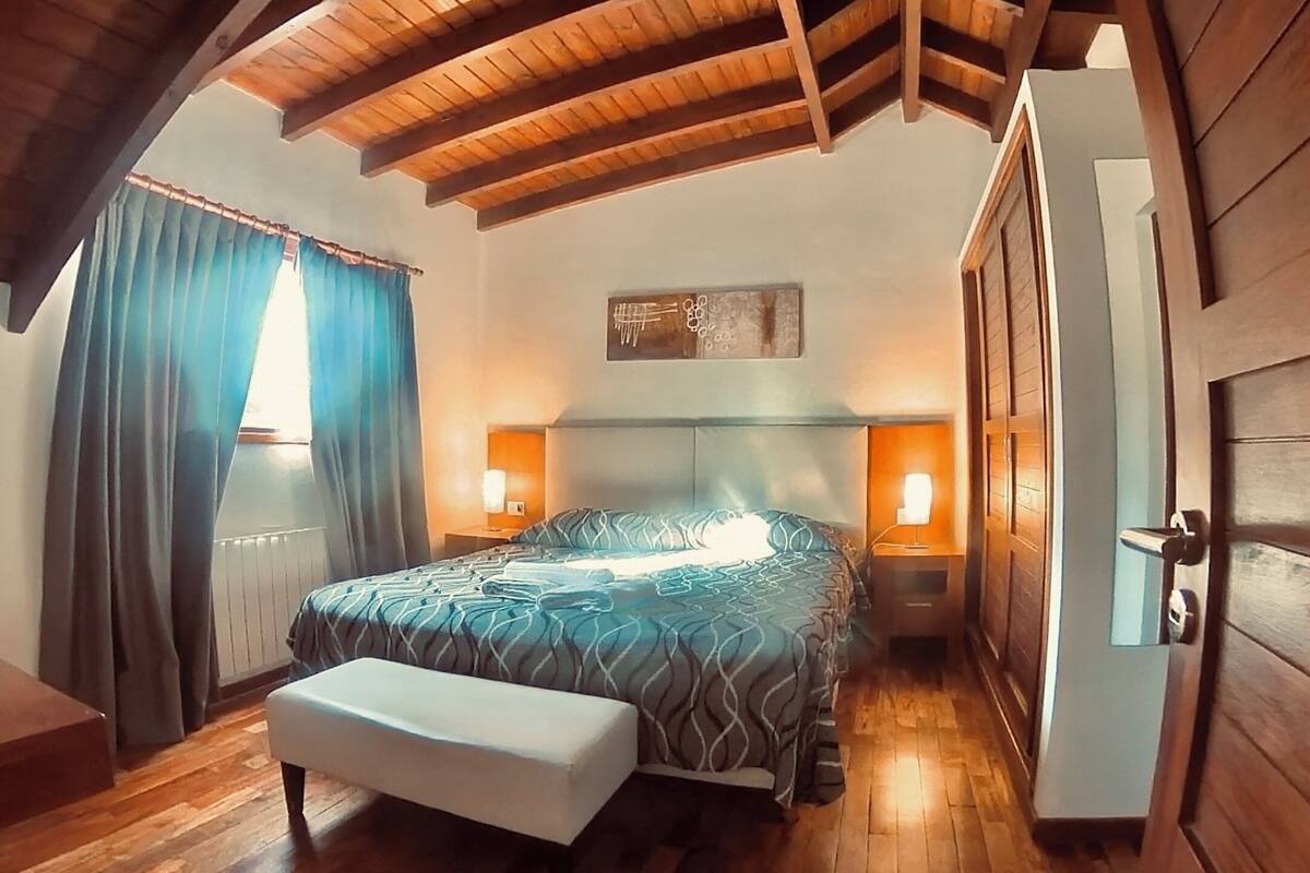 A bedroom with a wooden ceiling, a bed with patterned bedding, bedside lamps, and a window with curtains.