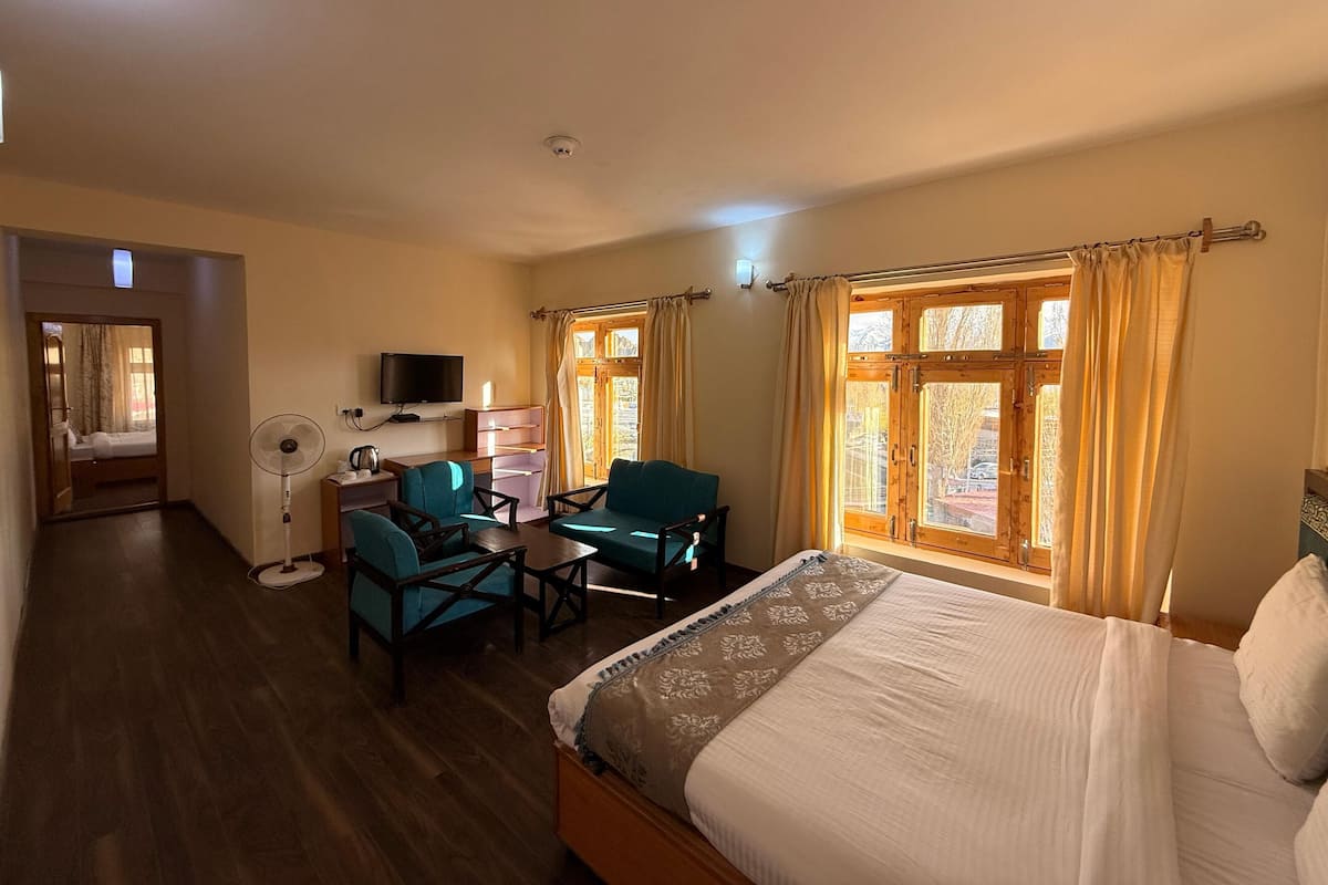 A hotel room with a bed, two chairs, a desk with a TV, a fan, and a window with curtains.