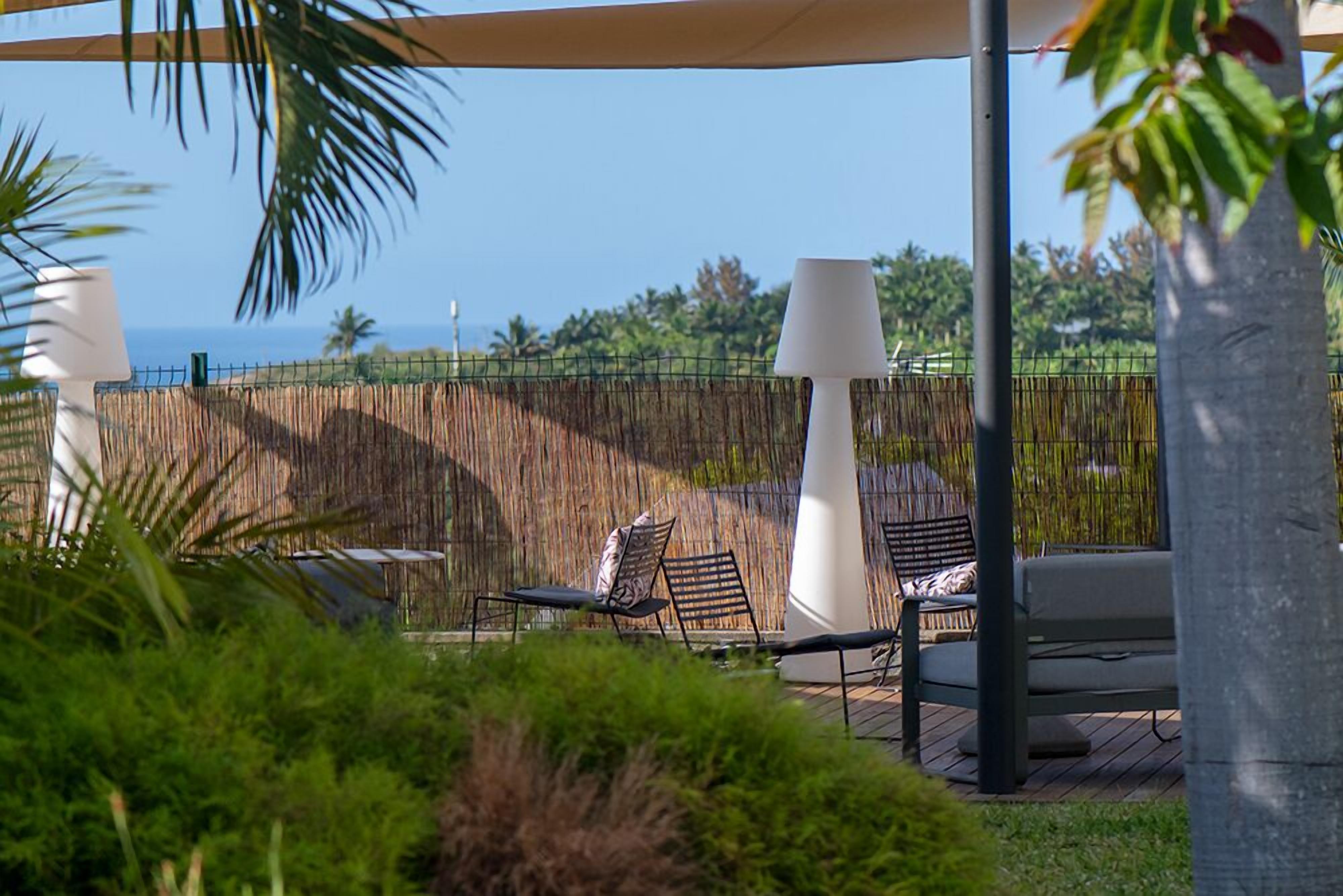 A serene outdoor seating area with lounge chairs and a table, surrounded by a bamboo fence and lush greenery, overlooking a clear blue sky and distant landscape.