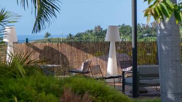 A serene outdoor seating area with lounge chairs and a table, surrounded by a bamboo fence and lush greenery, overlooking a clear blue sky and distant landscape.