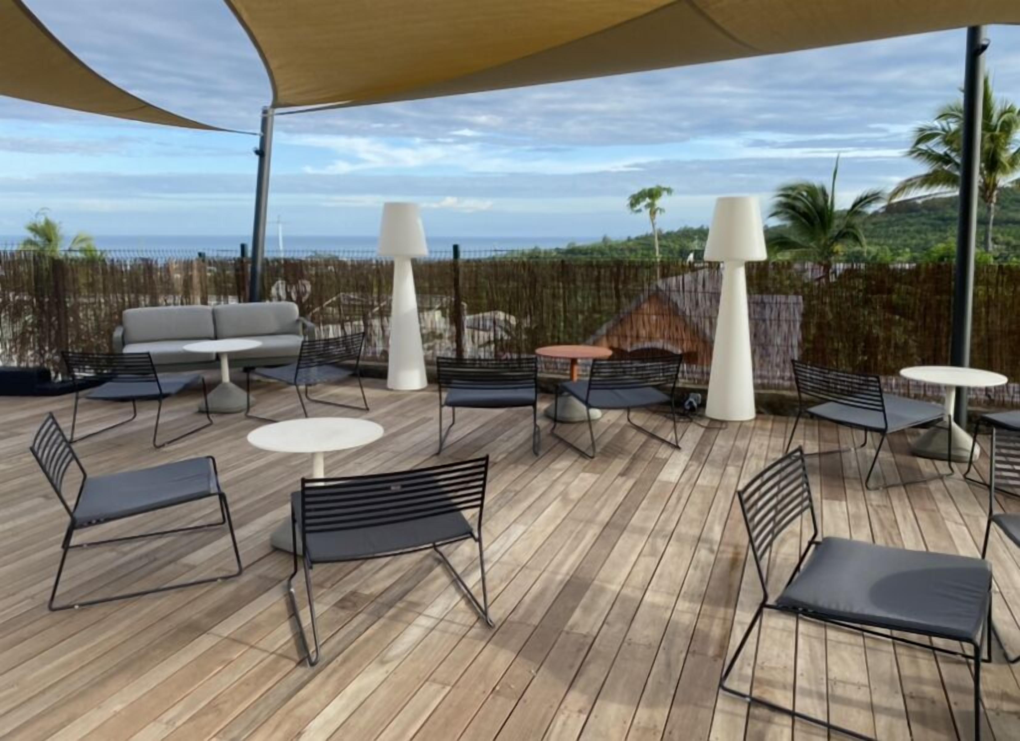 Een terras buiten met houten vloer, zwarte metalen stoelen, ronde tafels en een witte bank.
