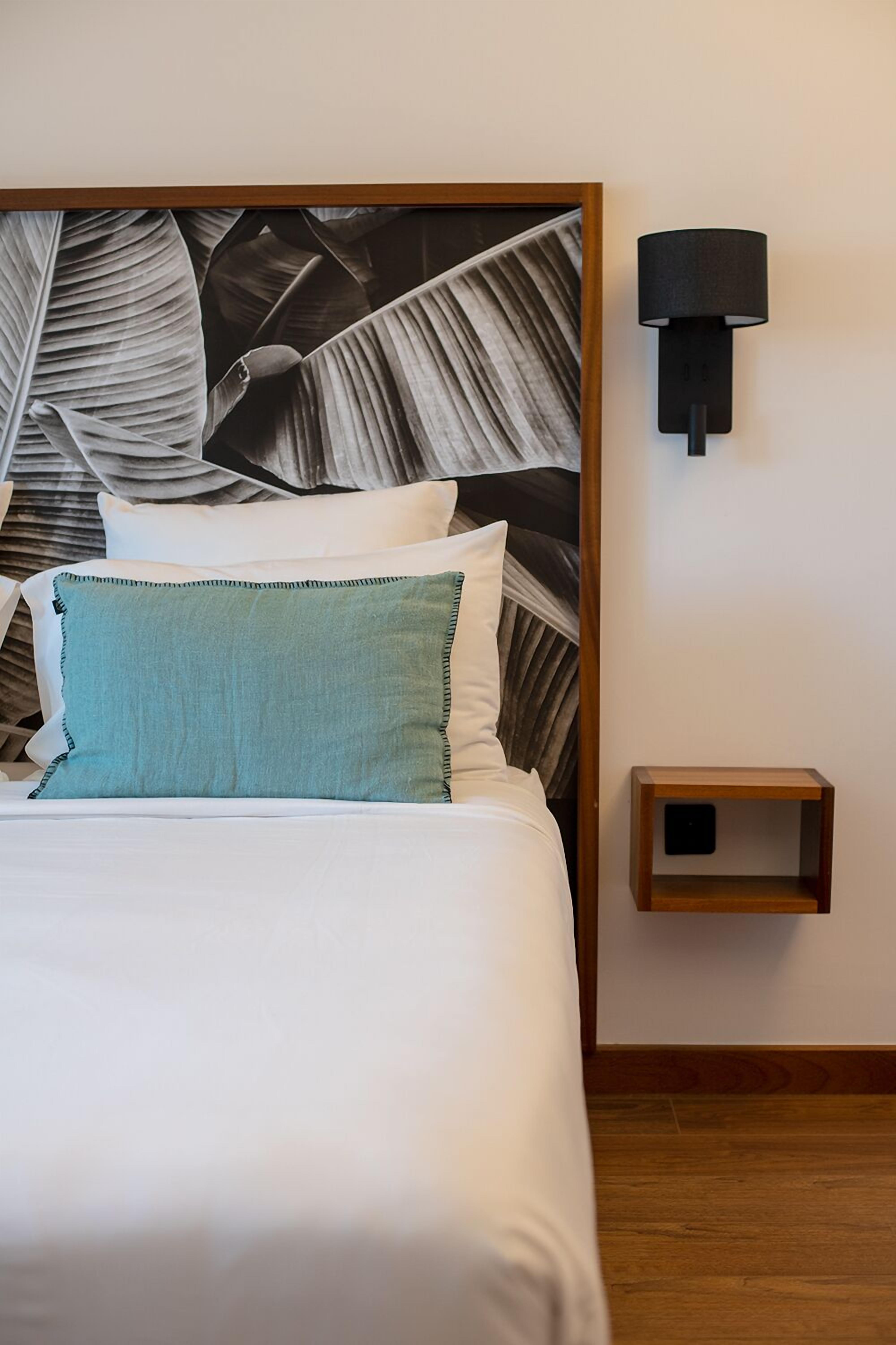 Una cama bien hecha con un almohá de color turquesa, una lámpara negra fijada a la pared y una mesita de noche de madera.