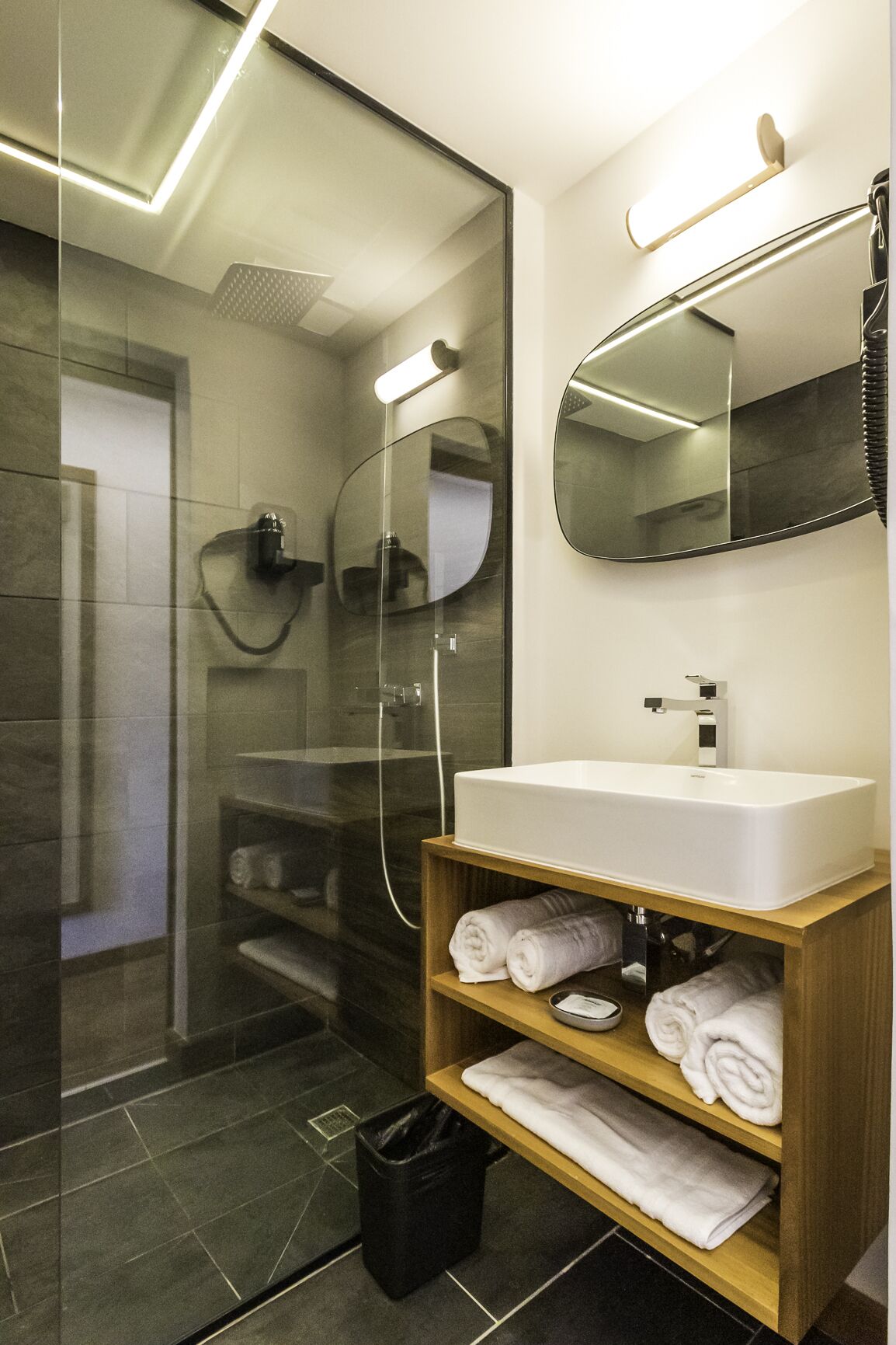 Un baño moderno con ducha de vidrio, un lavabo blanco y un mueble de tocador de madera con toallas.