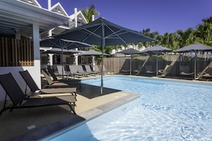 Una piscina con sillones de descanso y sombrillas, rodeada por una valla de madera y palmeras.