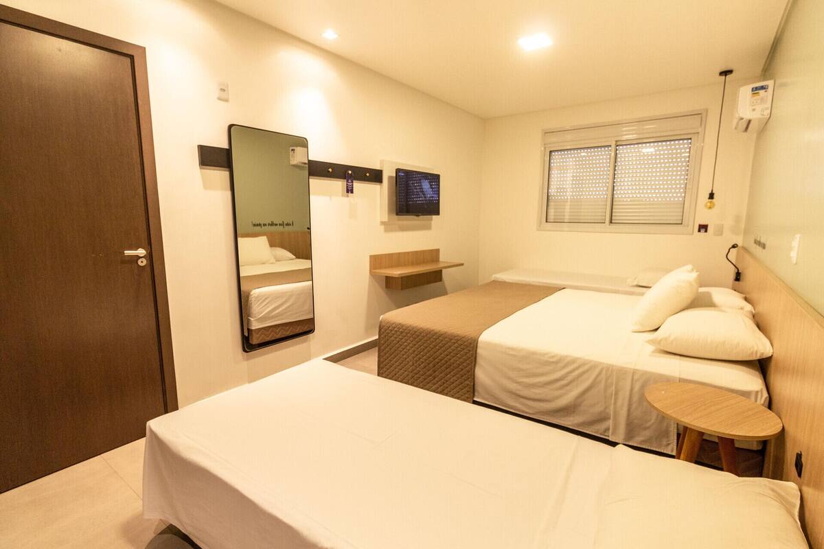 A hotel room with a bed, a bedside table, a mirror, a TV, and a window with blinds.