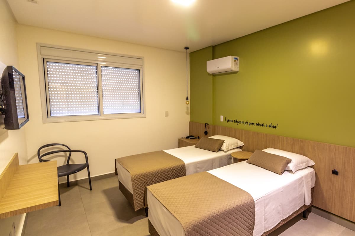 A hotel room with two beds, a desk, a chair, a window with blinds, an air conditioning unit, and a wall with a motivational quote.
