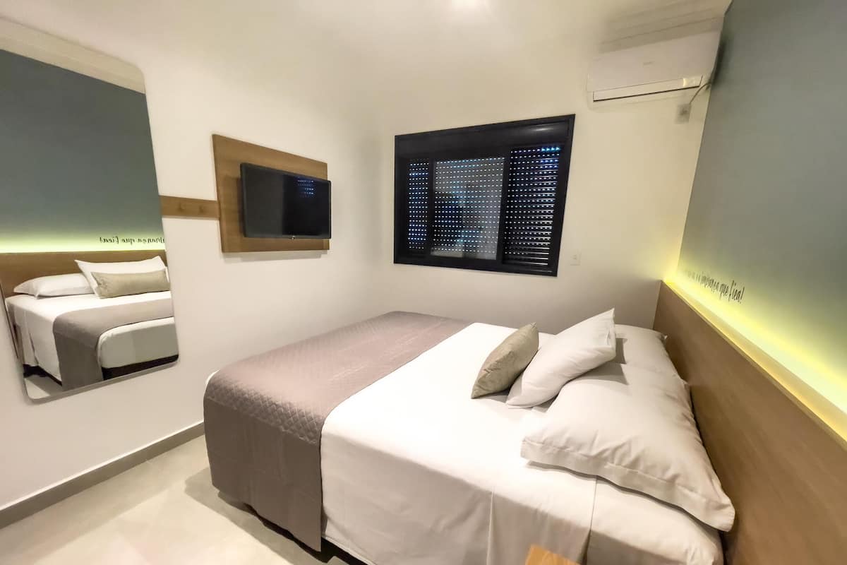 A modern hotel room with a bed, a TV, a window with blinds, and a wall-mounted air conditioner.