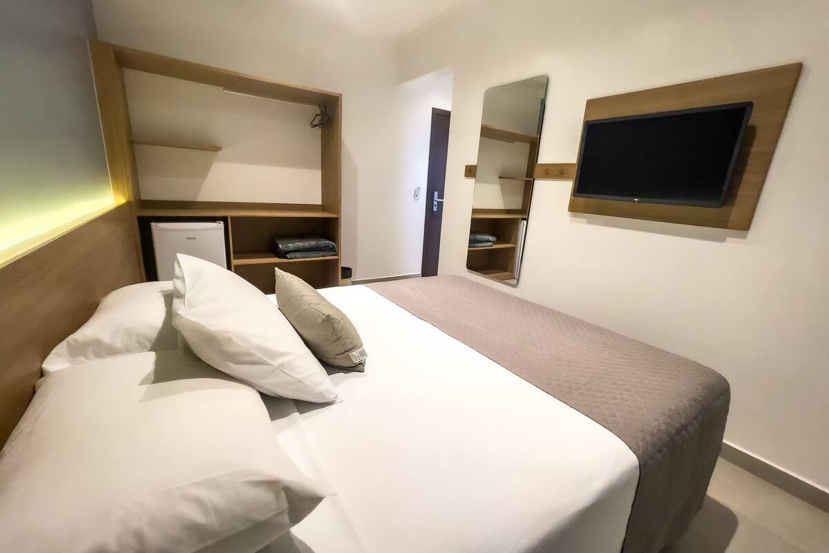 A neatly made bed with white and beige bedding, a wooden headboard, and a bedside table with a lamp. There is a built-in shelf with a mirror and a flat-screen TV mounted on the wall.