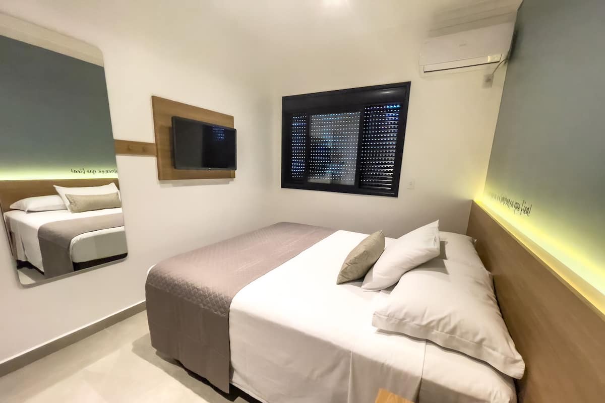 A modern hotel room with a large bed, a TV mounted on the wall, and a window with blinds.