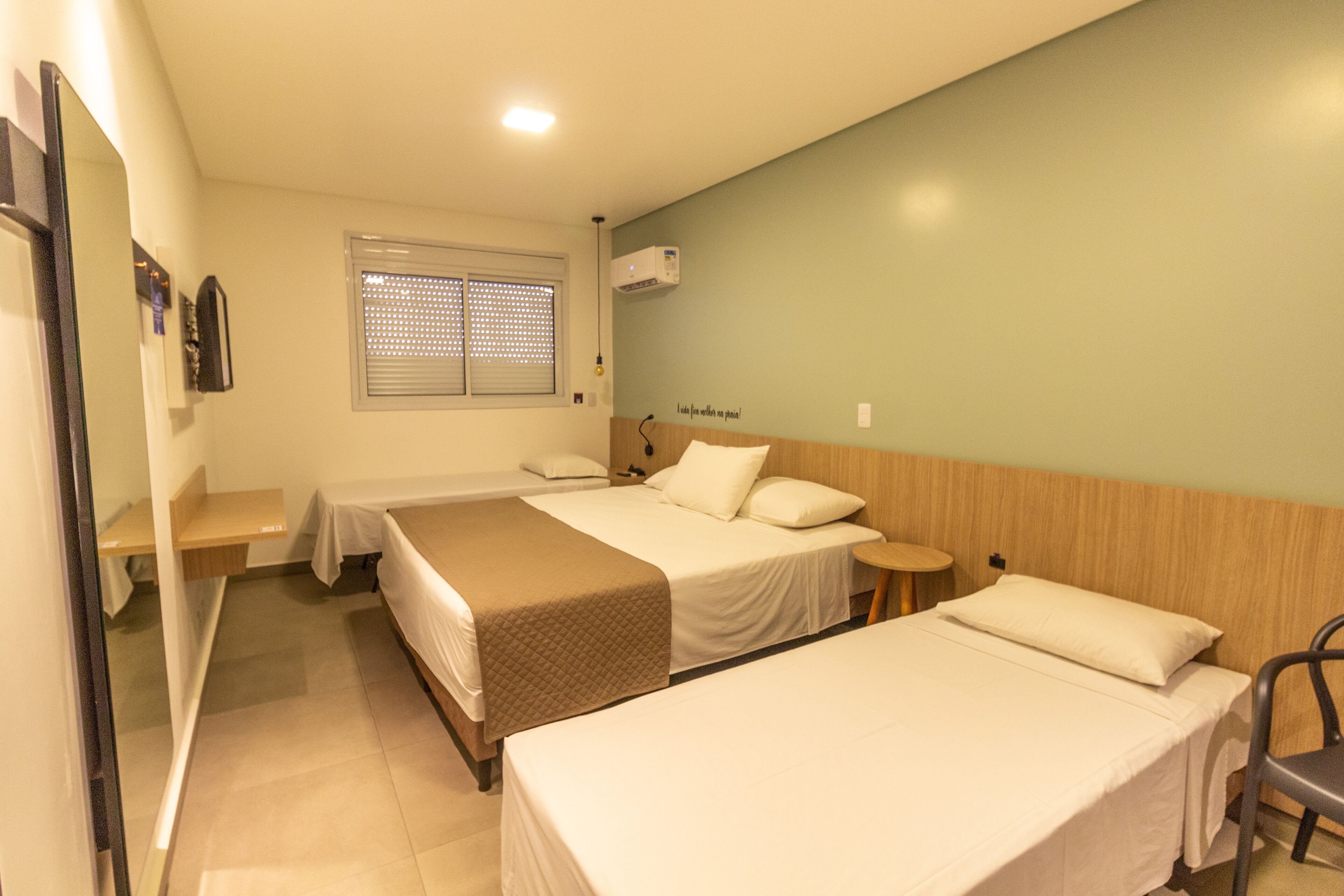 Habitación de hotel con dos camas individuales, una mesita, una silla, una ventana con persianas y un equipo de aire acondicionado en la pared.