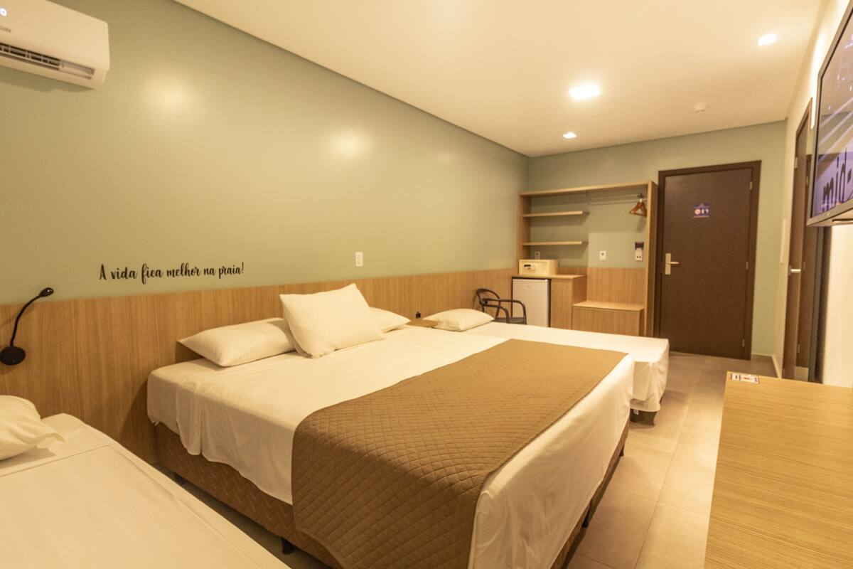 A hotel room with two beds, a wooden headboard, a wall-mounted air conditioner, and a shelf with a mirror and a hanging coat rack.