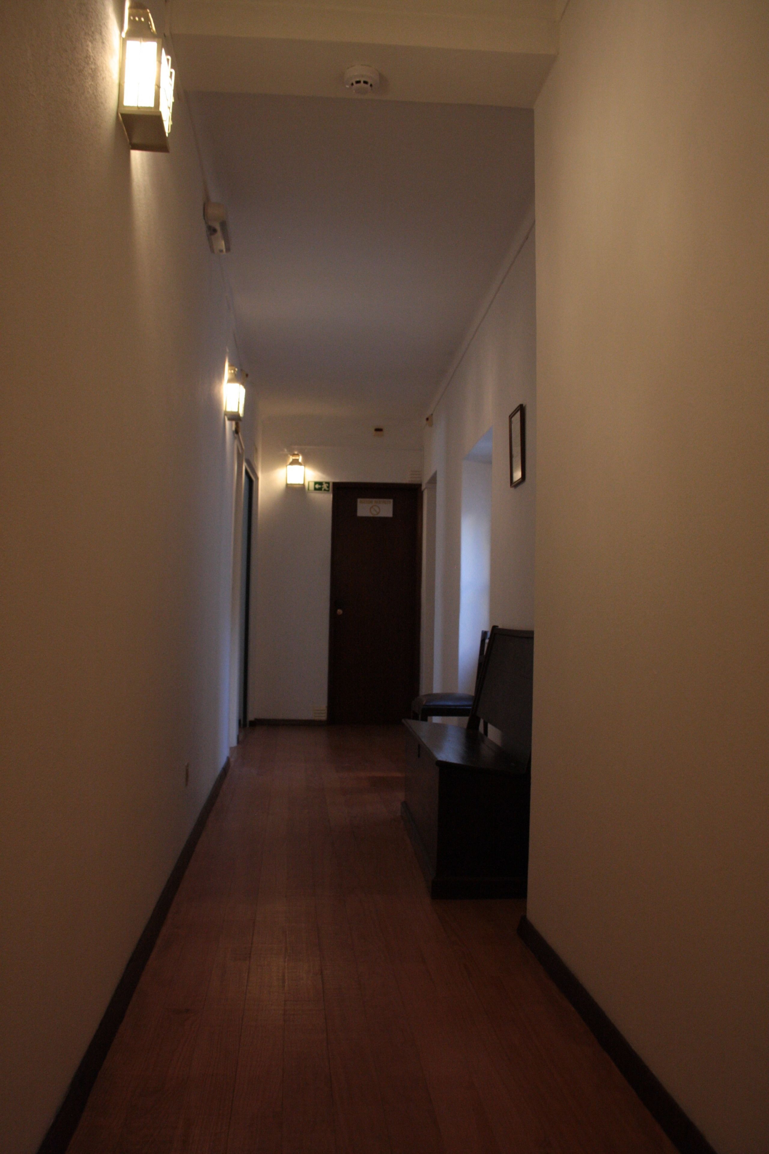 A narrow hallway with wooden flooring, a door at the end, and a bench on the right.