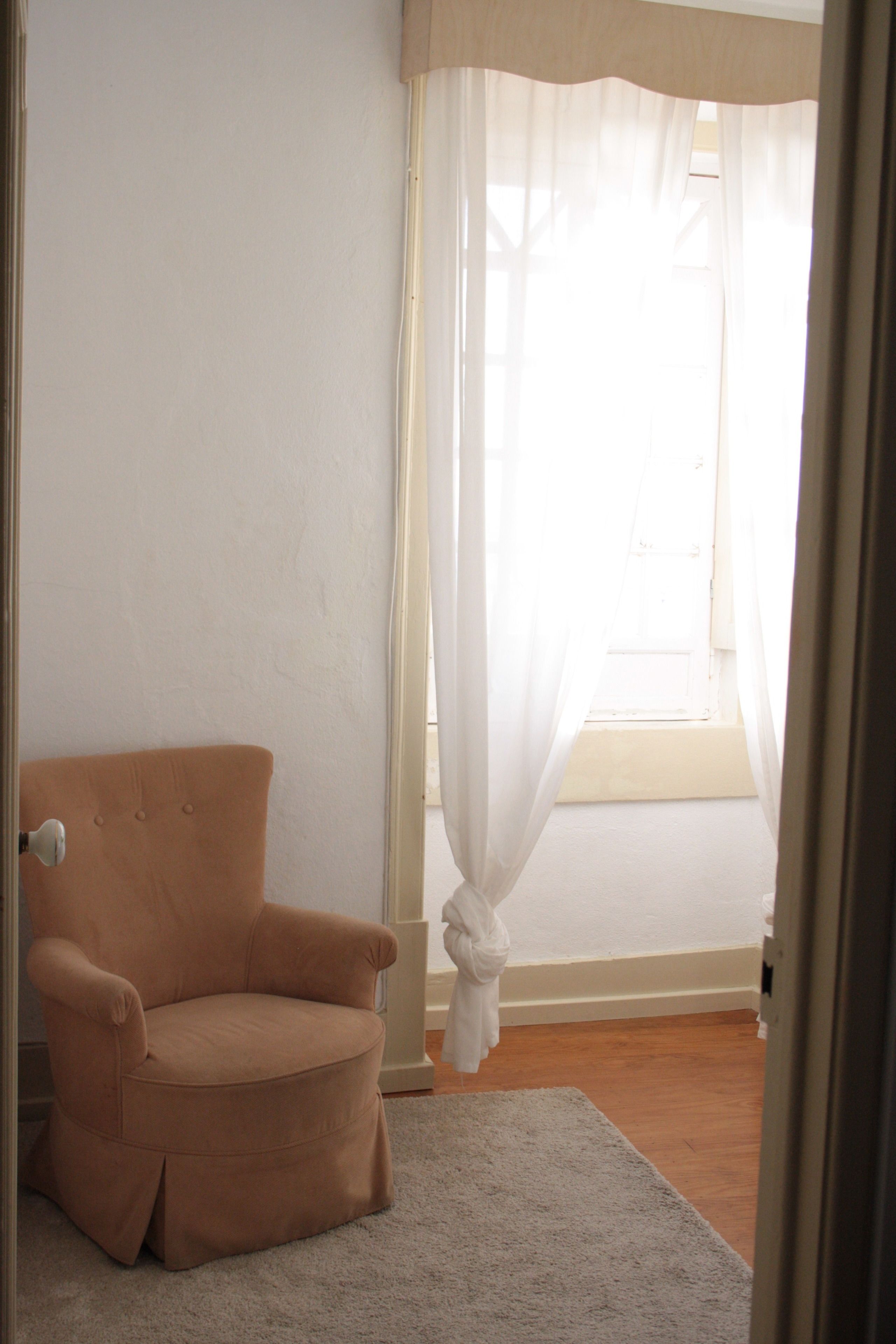 A room with a beige armchair, a white carpet, and a window with sheer curtains.