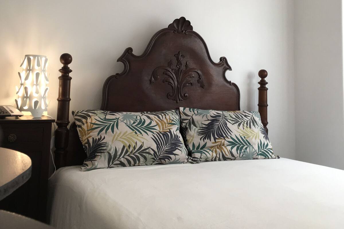 A bedroom with a wooden headboard, a bed with a leaf-patterned pillow, a bedside table with a lamp, and a wall-mounted light fixture.