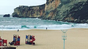 A beach with a rocky cliff, waves, and a lifeguard tower. - Hotel Ribamar (Nazaré)