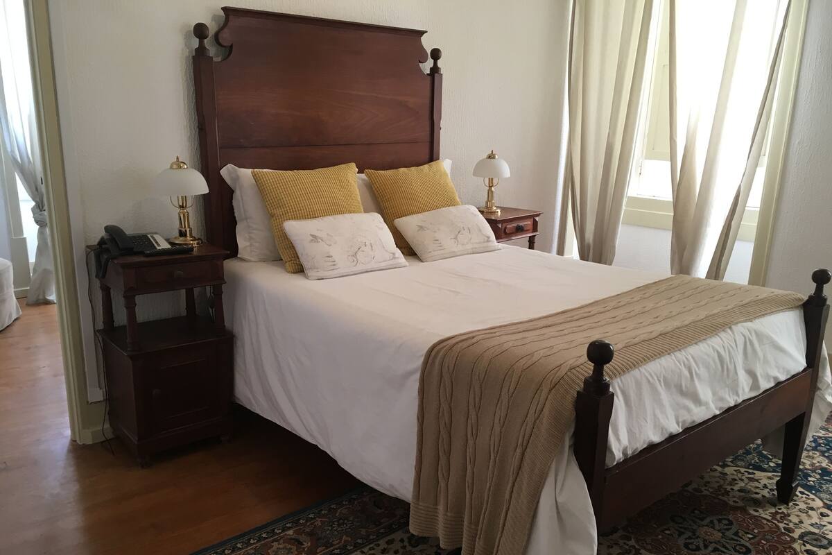 A neatly made bed with a wooden headboard, white bedding, and two decorative pillows. There is a bedside table with a lamp and a phone. The room has a patterned rug and a window with curtains.