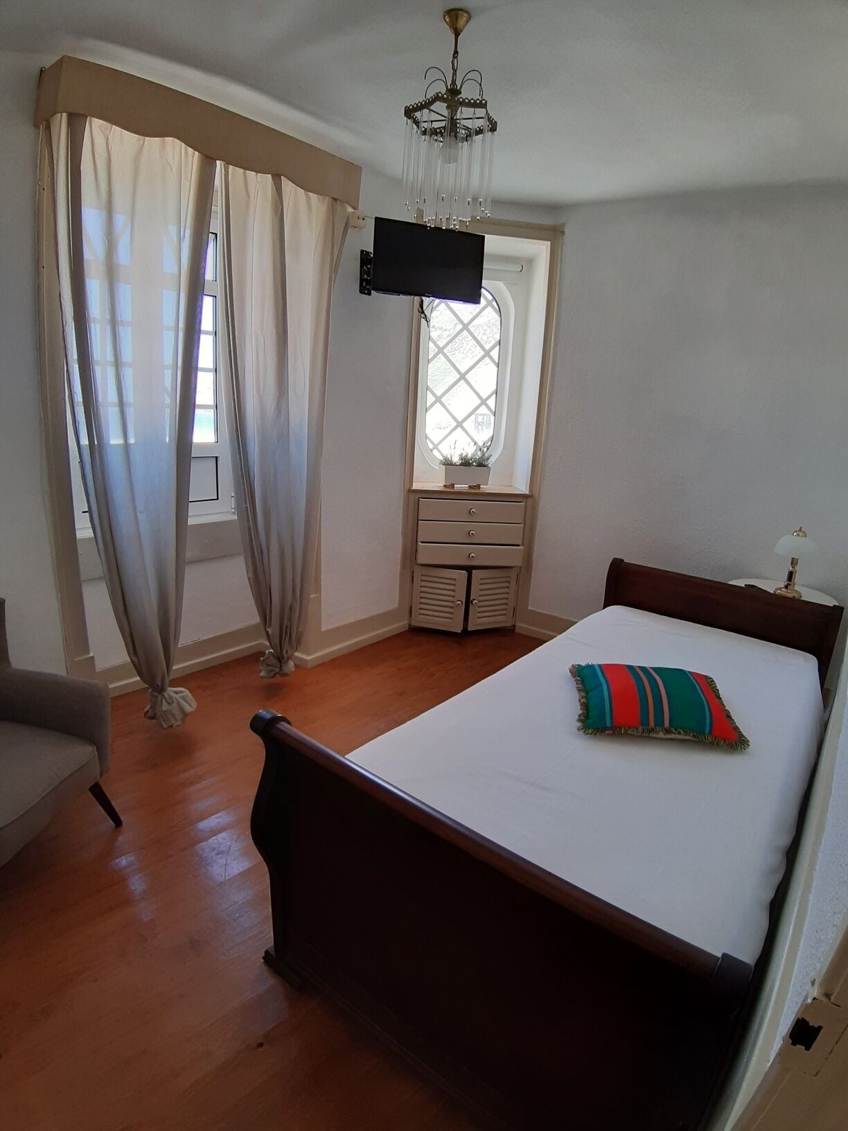 Ein Schlafzimmer mit einem hölzernen Bett, einem kleinen Nachttisch, einem Stuhl, einem Fenster mit Vorhängen und einer Deckenleuchte.