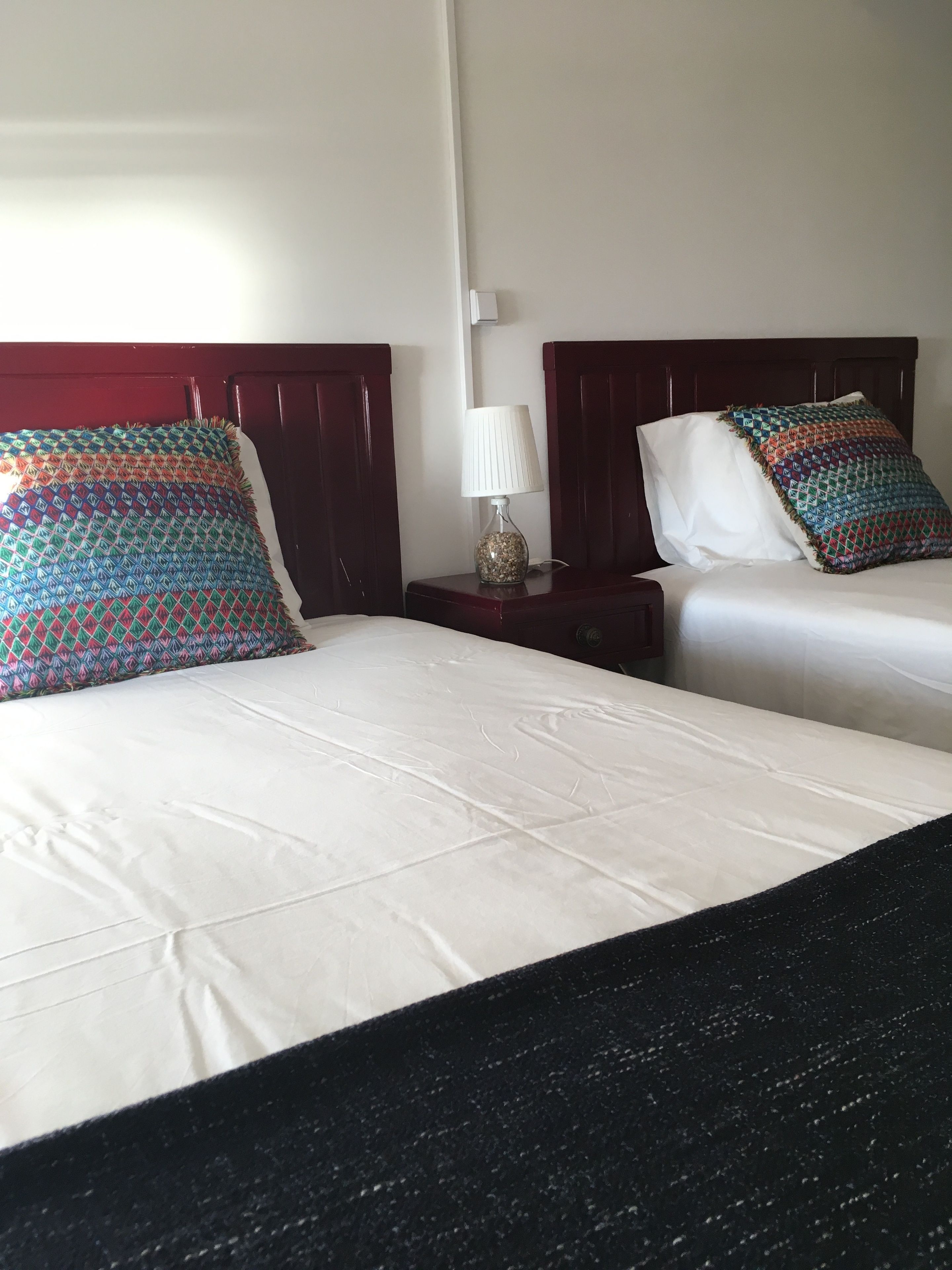 Habitación de hotel con dos camas, cada una con un cojín estampado y sábanas blancas. Hay una mesita de noche con una lámpara entre las camas.