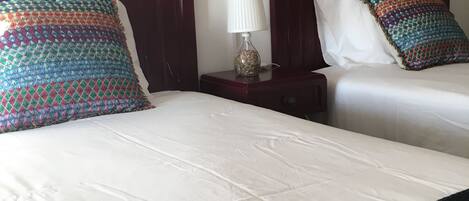 Habitación de hotel con dos camas, cada una con un cojín estampado y sábanas blancas. Hay una mesita de noche con una lámpara entre las camas.