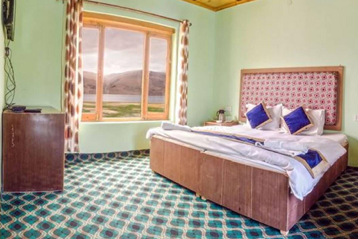 Un dormitorio con cama, cabecera de madera, ventana con cortinas, suelo estampado y un escritorio.