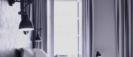 A modern hotel room with a neatly made bed, a nightstand with a lamp, and a view of the outdoors through a large window.
