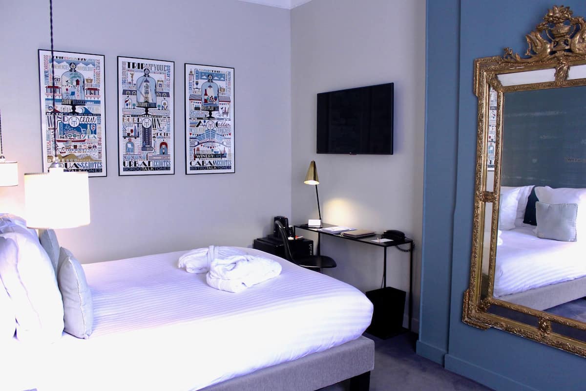 Quarto de hotel com uma cama bem arrumada, uma escrivaninha com luminária, um espelho grande e três quadros na parede.