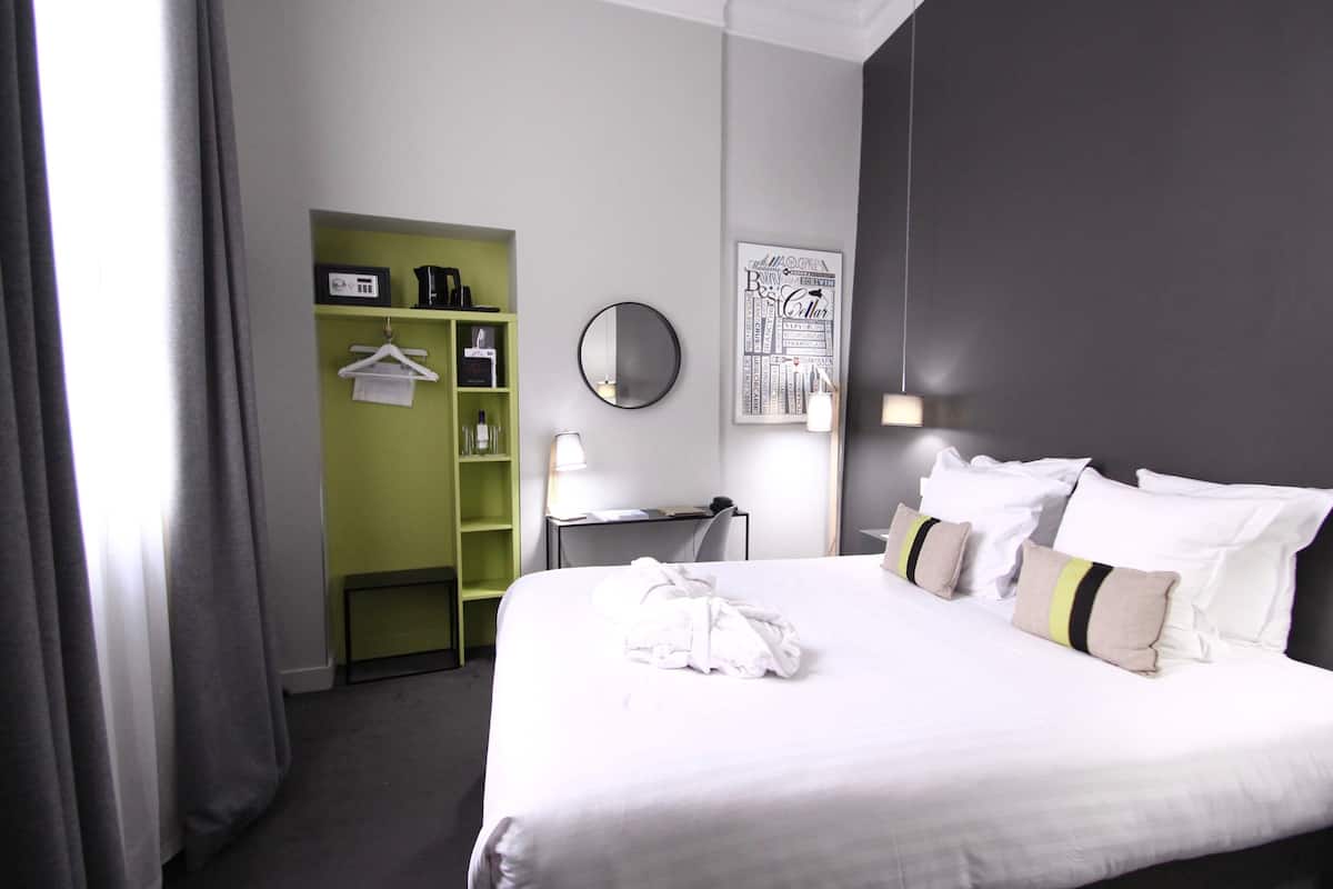 Quarto de hotel com cama, escrivaninha, espelho, guarda-roupa verde e uma obra de arte na parede.