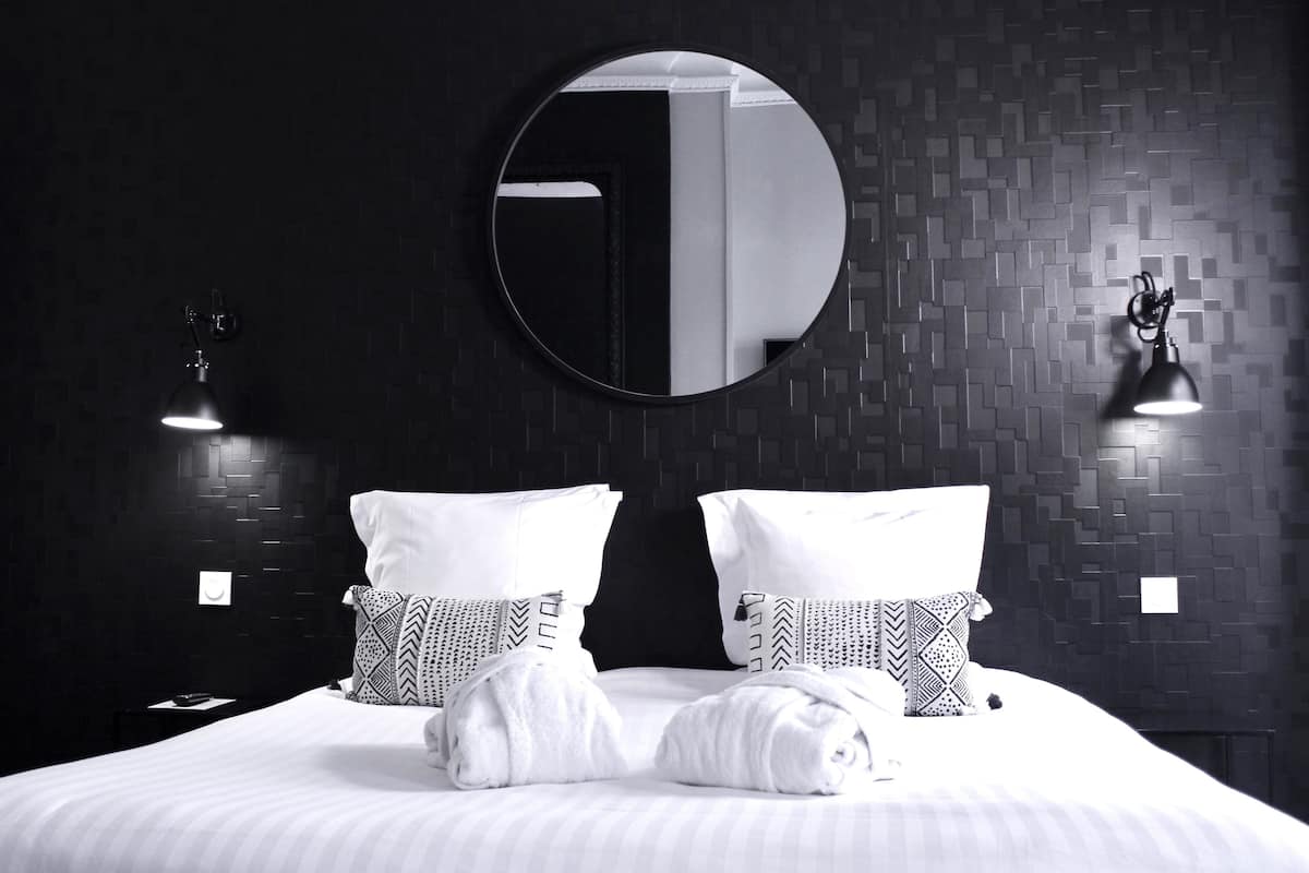 Quarto de hotel com uma cama grande, duas almofadas estampadas, um espelho redondo e duas luminárias fixadas na parede.