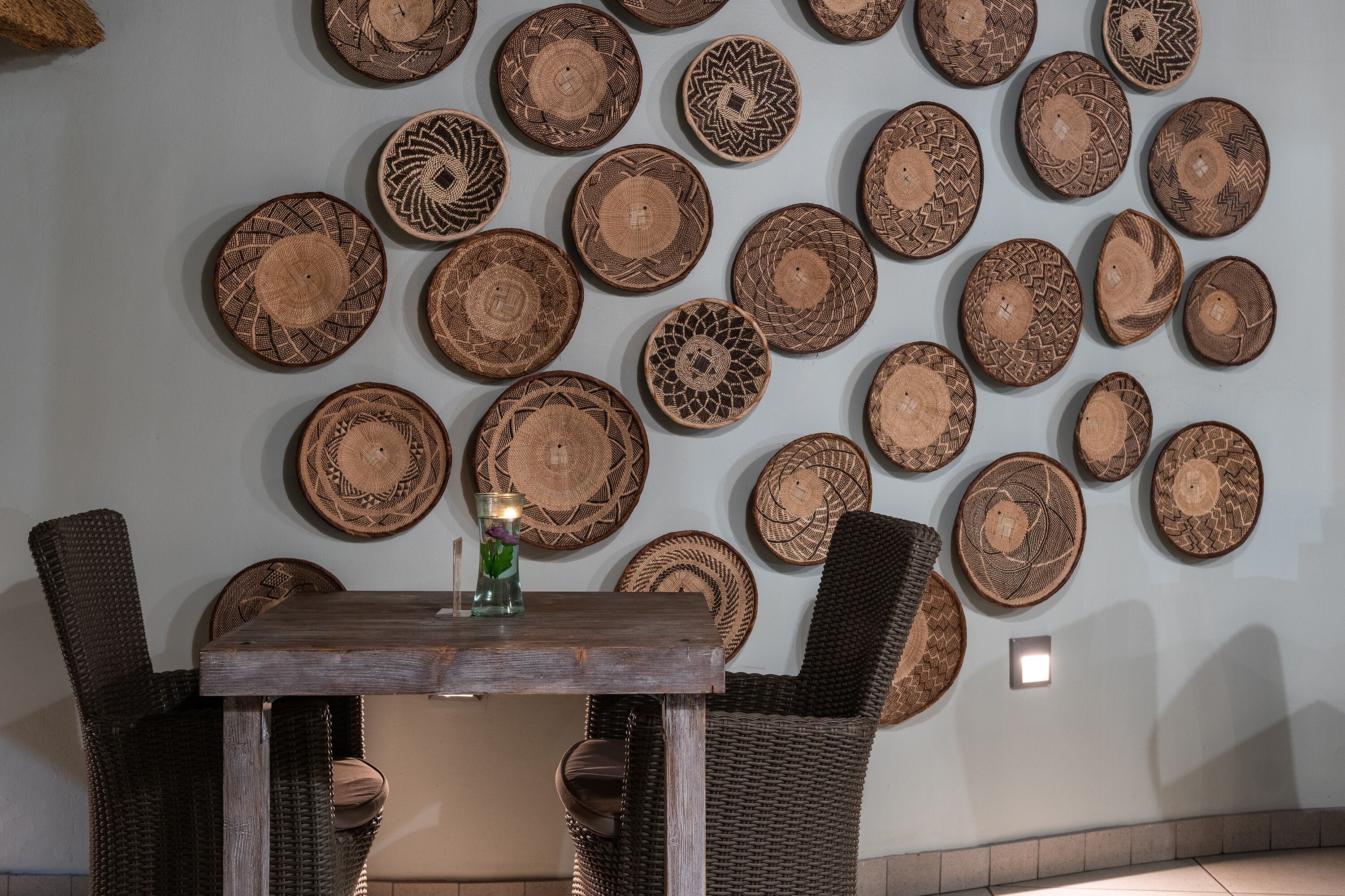 A dining area with a wooden table and wicker chairs, surrounded by a wall of woven plates.