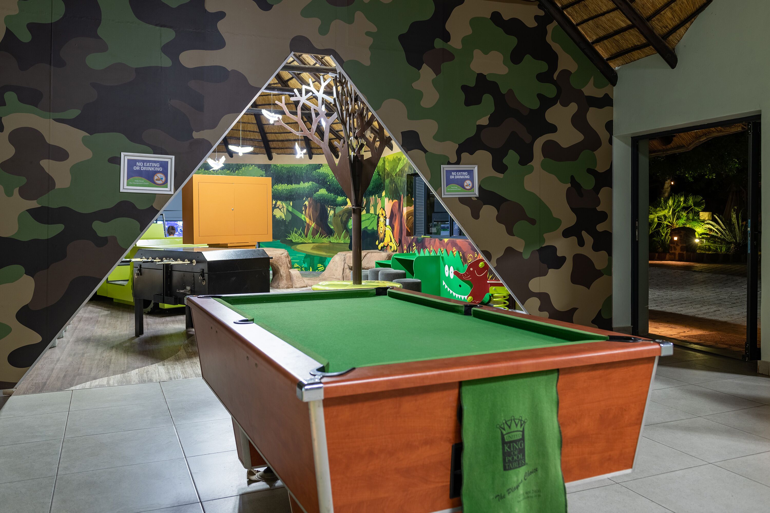 A pool table with a green felt surface, surrounded by camouflage-patterned walls and a mural depicting a jungle scene.