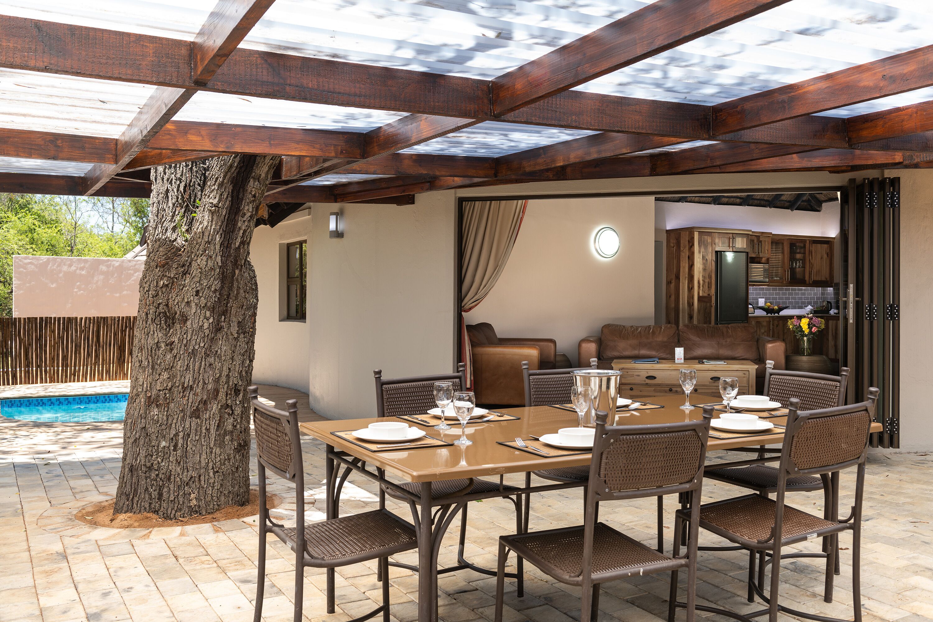 Un espacio de comedor al aire libre con mesa y sillas de madera, piscina y cocina visible a través de un umbral abierto.