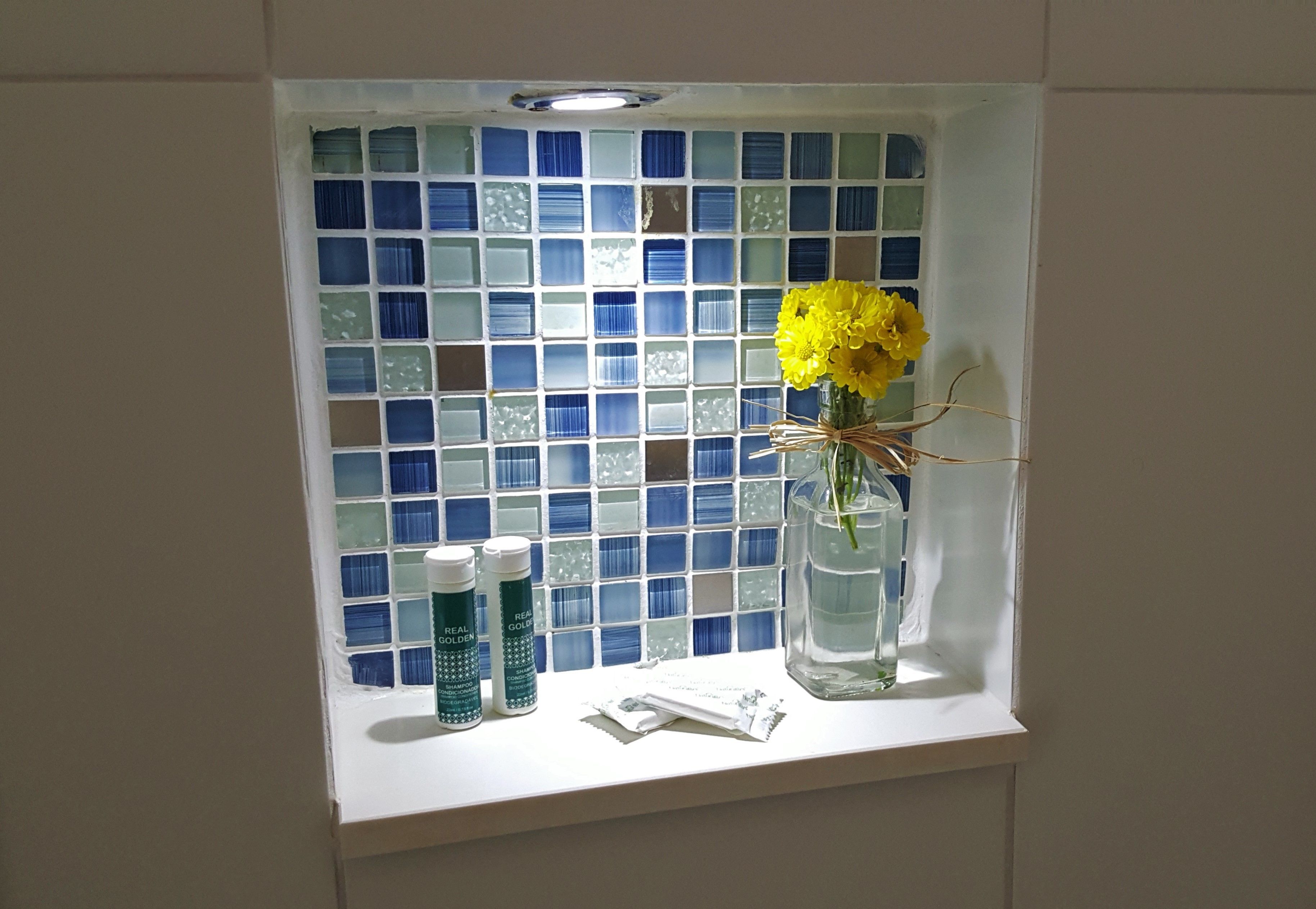 Un tocador de baño con un salpicadero de mosaicos azules, un florero transparente con flores amarillas y dos botellas de loción.