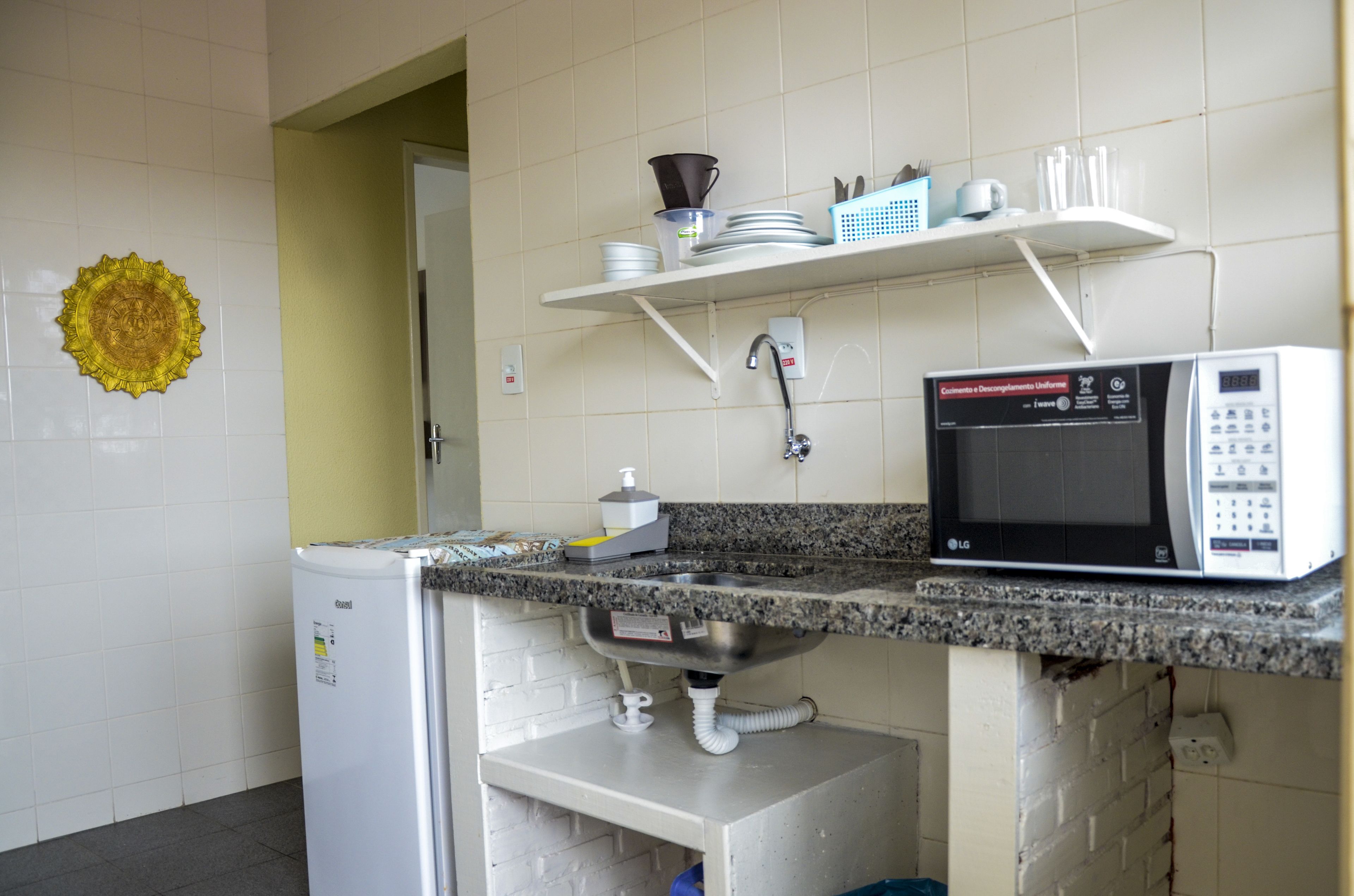 A compact kitchen with a built-in microwave, a small refrigerator, and a shelf with kitchenware.