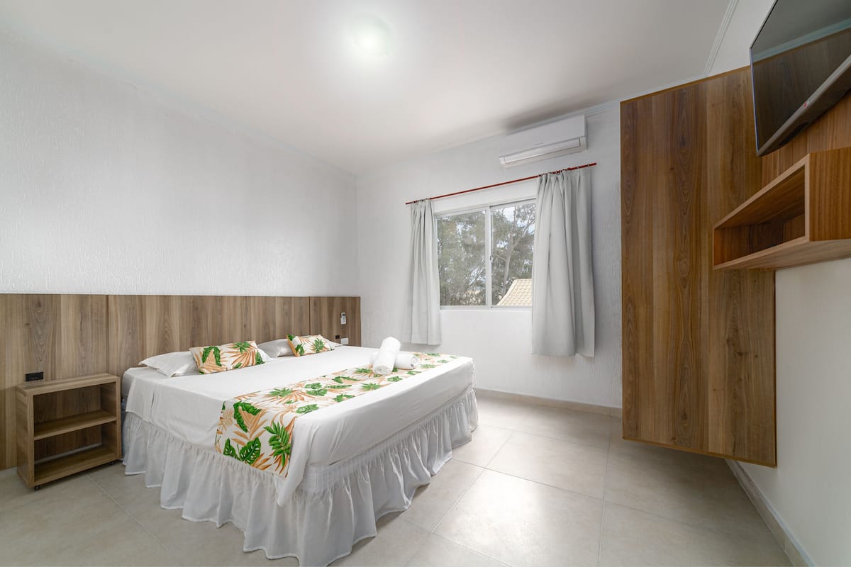 A bedroom with a bed, a wooden headboard, a TV mounted on the wall, and a window with curtains.