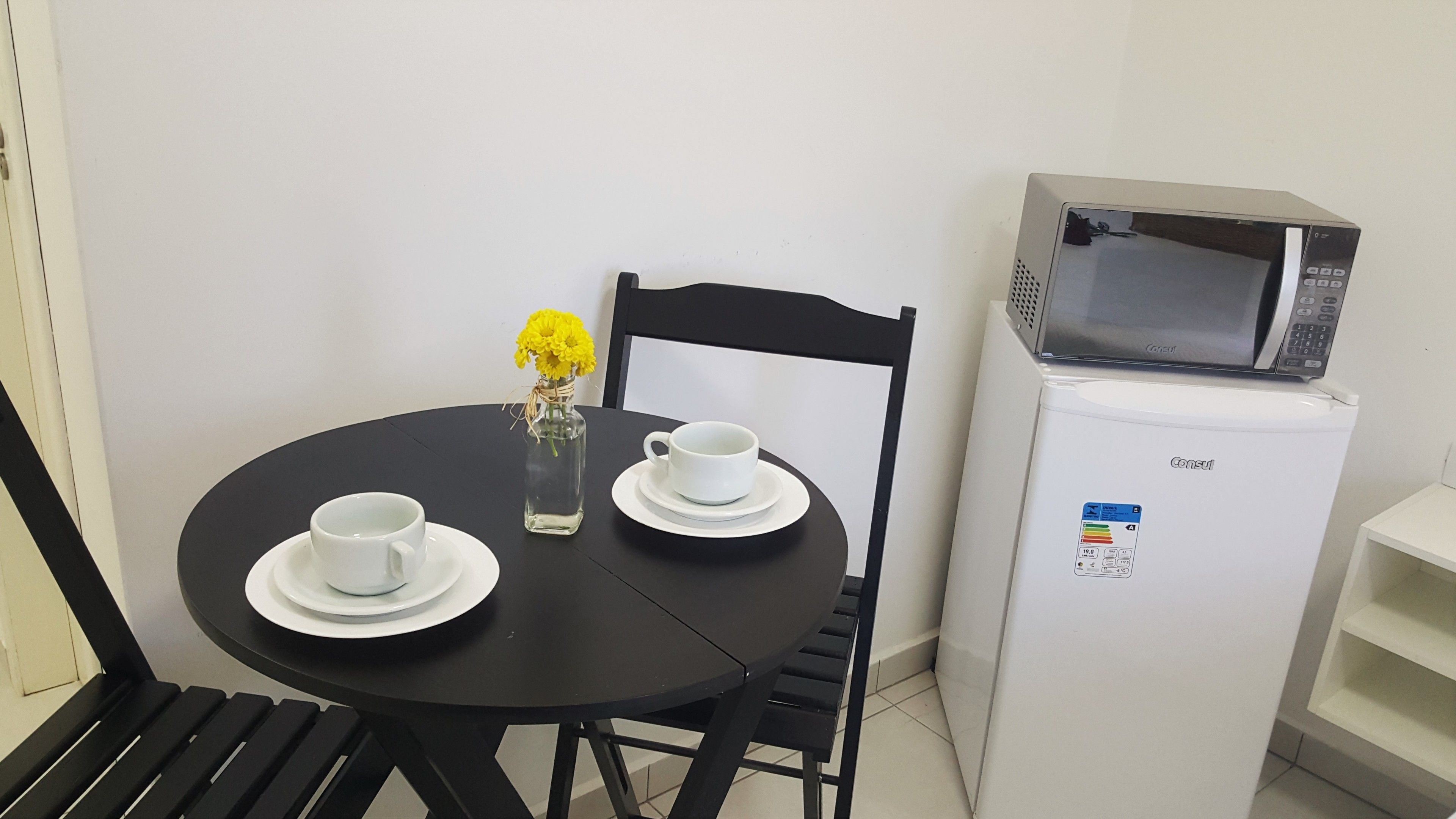 A small dining area with a black table, two white cups on saucers, a clear glass vase with yellow flowers, a black chair, a white refrigerator with a microwave, and a white shelf.